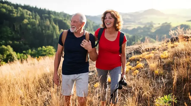 Ein älteres Paar wandert im Sommer über das Land, im Hintergrund sind bewaldete Hügel zu sehen.