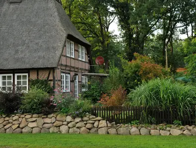 Ferienhaus in Niedersachsen Fachwerkhaus mit Krüppelwalmdach ist mit Schilf gedeckt und umgeben von grünen Büschen