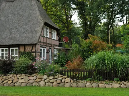 Fachwerkhaus mit Krüppelwalmdach ist mit Schilf gedeckt und umgeben von grünen Büschen