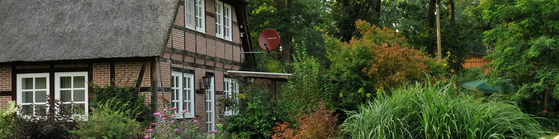 Fachwerkhaus mit Krüppelwalmdach ist mit Schilf gedeckt und umgeben von grünen Büschen