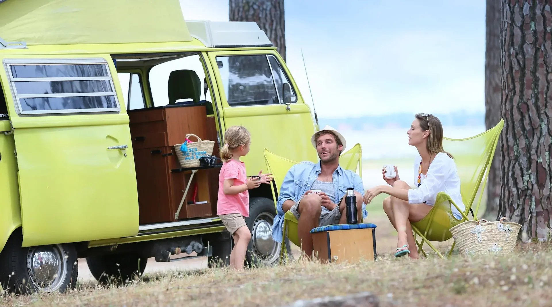Ein grüner, nostalgischer Campingbus steht auf sandigem Boden unter schattigen Bäumen. Davor sitzt eine Familie entspannt in Campingstühlen und genießt die Natur. 