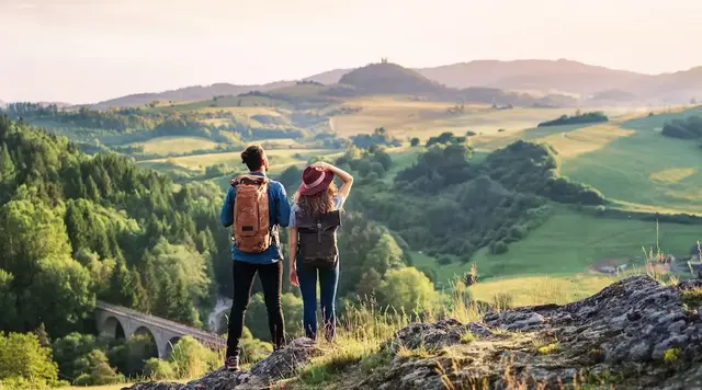 Ein junges Paar in Wanderkleidung und mit Rucksäcken steht vor einer weiten, hügeligen Landschaft. 