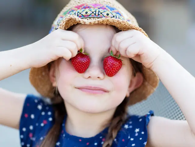 Erdbeeren selber pflücken – Spaß für Kinder auf dem Obsthof - © Anna Ritter, Adobe Stock Kind mit Sonnenhut hält sich zwei Erdbeeren vor die Augen auf einem Erdbeerfeld während eines Urlaubs auf dem Obsthof.