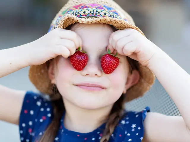 Kind mit Sonnenhut hält sich zwei Erdbeeren vor die Augen auf einem Erdbeerfeld während eines Urlaubs auf dem Obsthof.