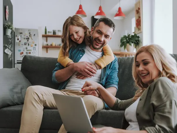 Happy family watching videos on laptop at home Cute little girl hugging her father from behind while he sitting on couch and holding hand of his wife. They looking at notebook display and smiling