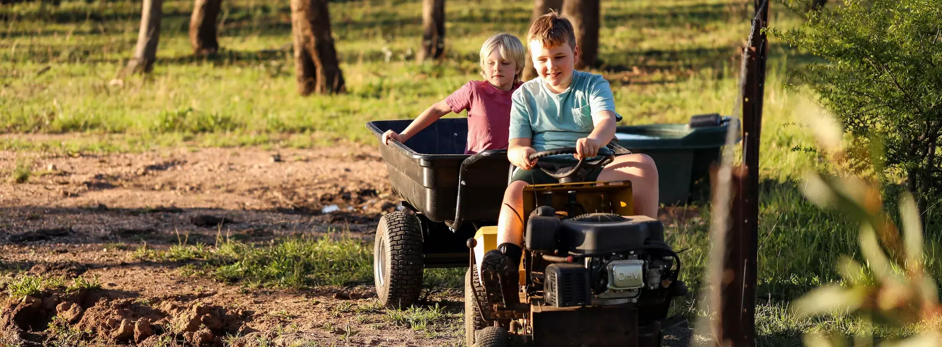 Zwei Jungs die glücklich auf einem Tretttraktor fahren. Der zweite Junge lässt sich in einem Anhänger durch die Natur ziehen.