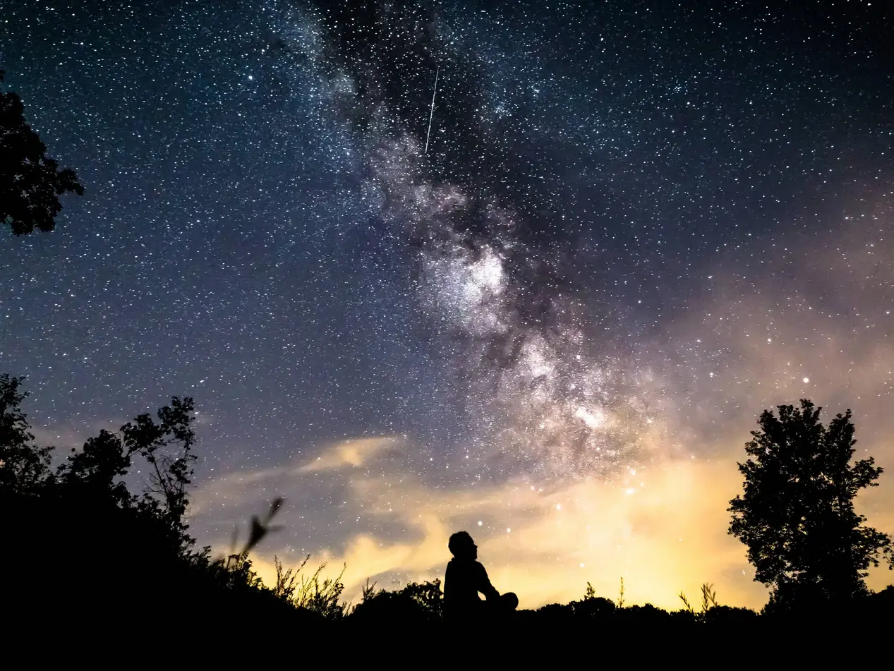 Mann unter Sternenhimmel in nächtlicher Landschaft - © egon999, adobe stock Mann sitzt nachts in einer Landschaft und blickt in den klaren Sternenhimmel.